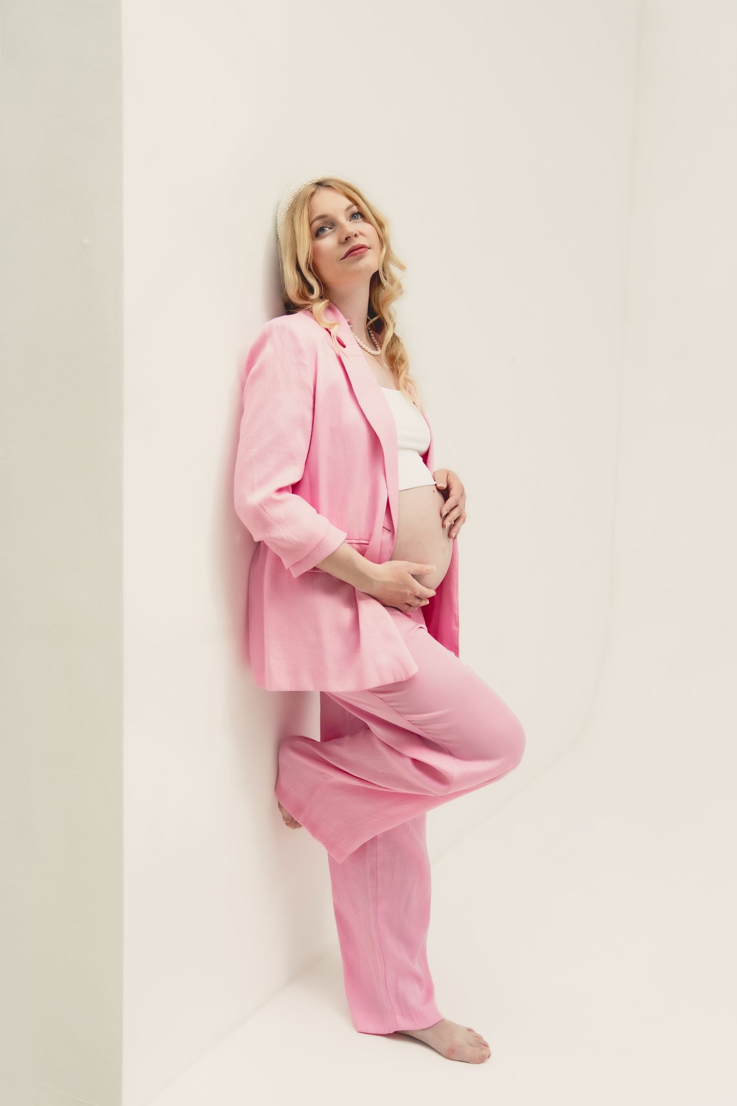 Pregnant woman in pink blazer and pants suit leaning against white wall, cradling baby bump.