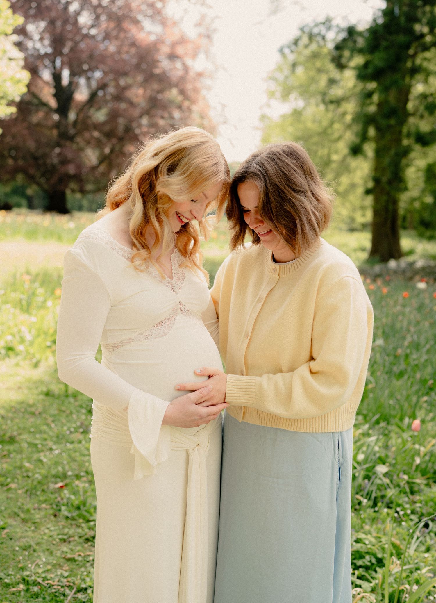 Two women smiling and cradling a baby bump in a sunlit garden with colorful wildflowers and lush green trees.