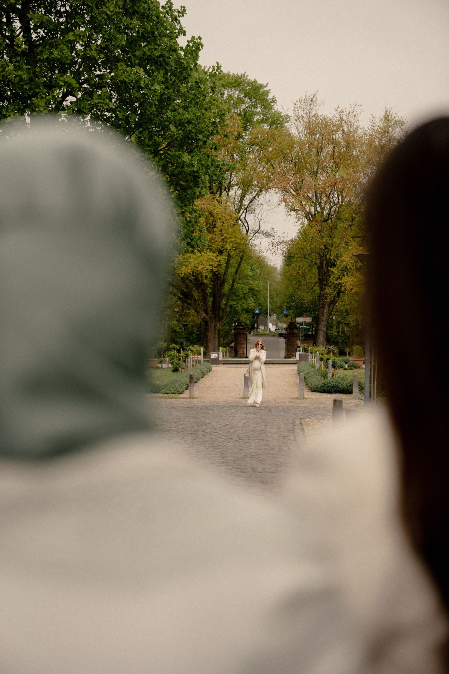 Bride in white dress standing in a tree-lined garden path viewed from behind two guests.