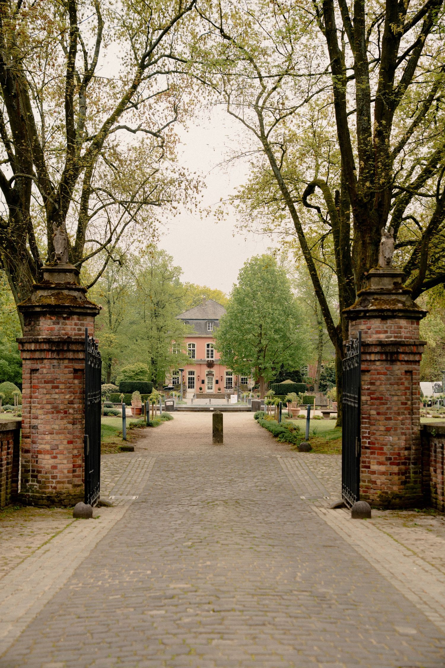 Elegant brick gate entrance leading to a charming historic manor house along a cobblestone path lined with tall trees.