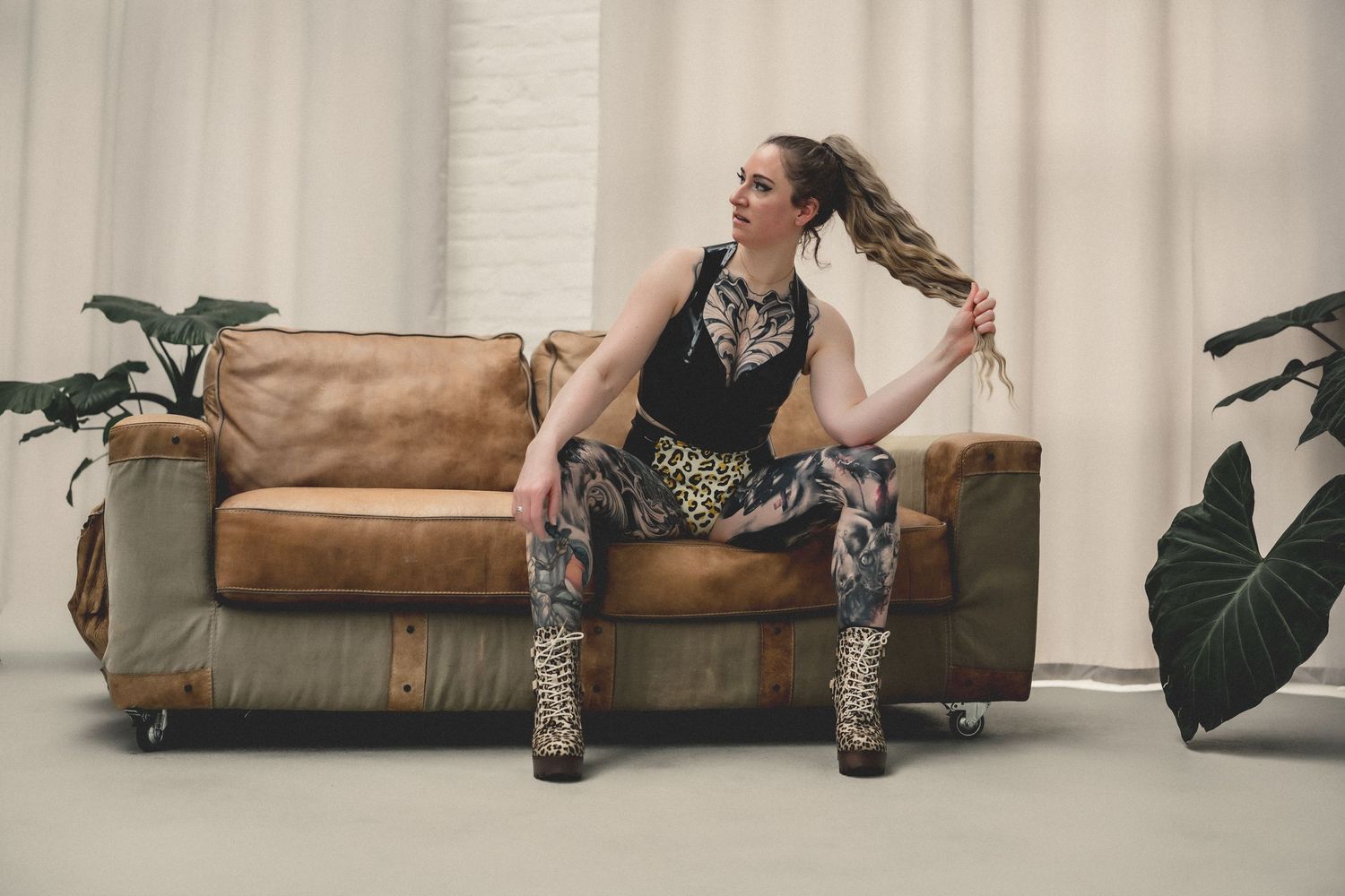 Woman in leopard print outfit sits on vintage leather sofa, posing with hair, flanked by tropical plants.