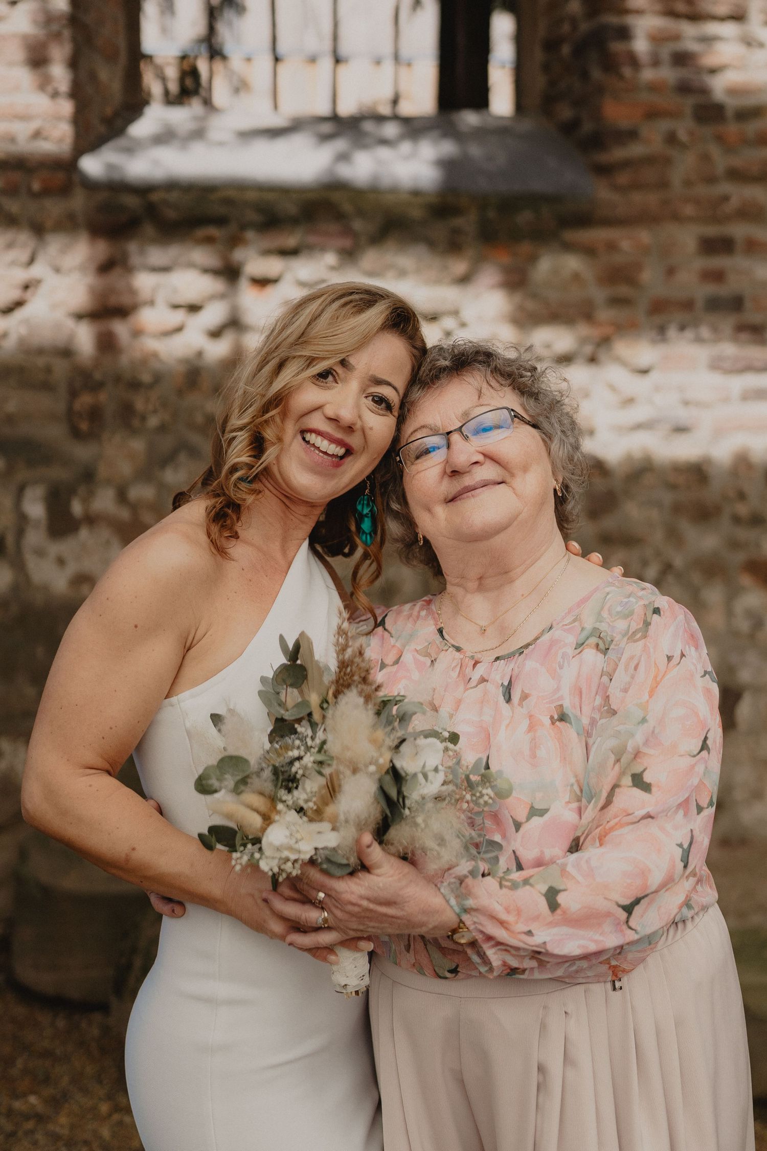 Tender emotional moment shared between two women in white and floral attire at a brick-walled venue.
