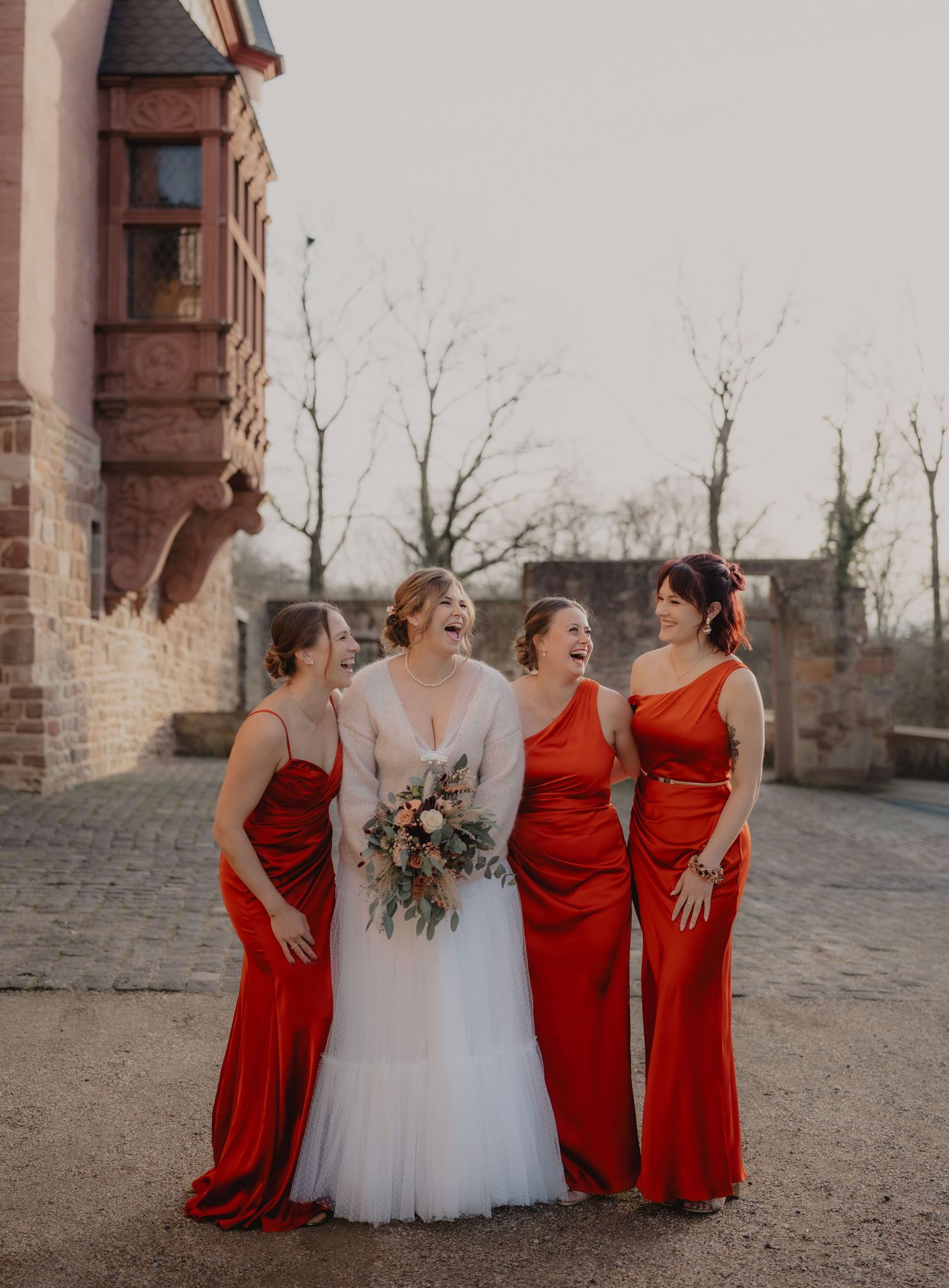 Wedding party in red bridesmaid dresses and white gown pose together by brick building.