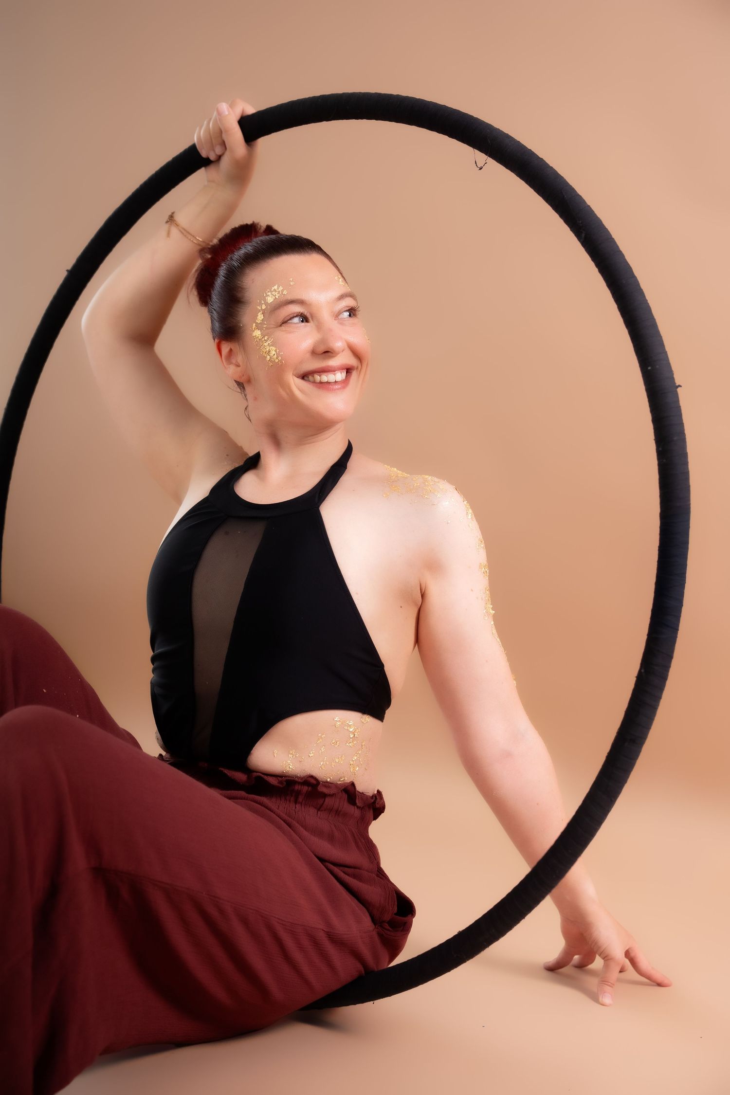 Smiling woman sitting with a large black hula hoop, wearing a black crop top and maroon pants with gold glitter accents.
