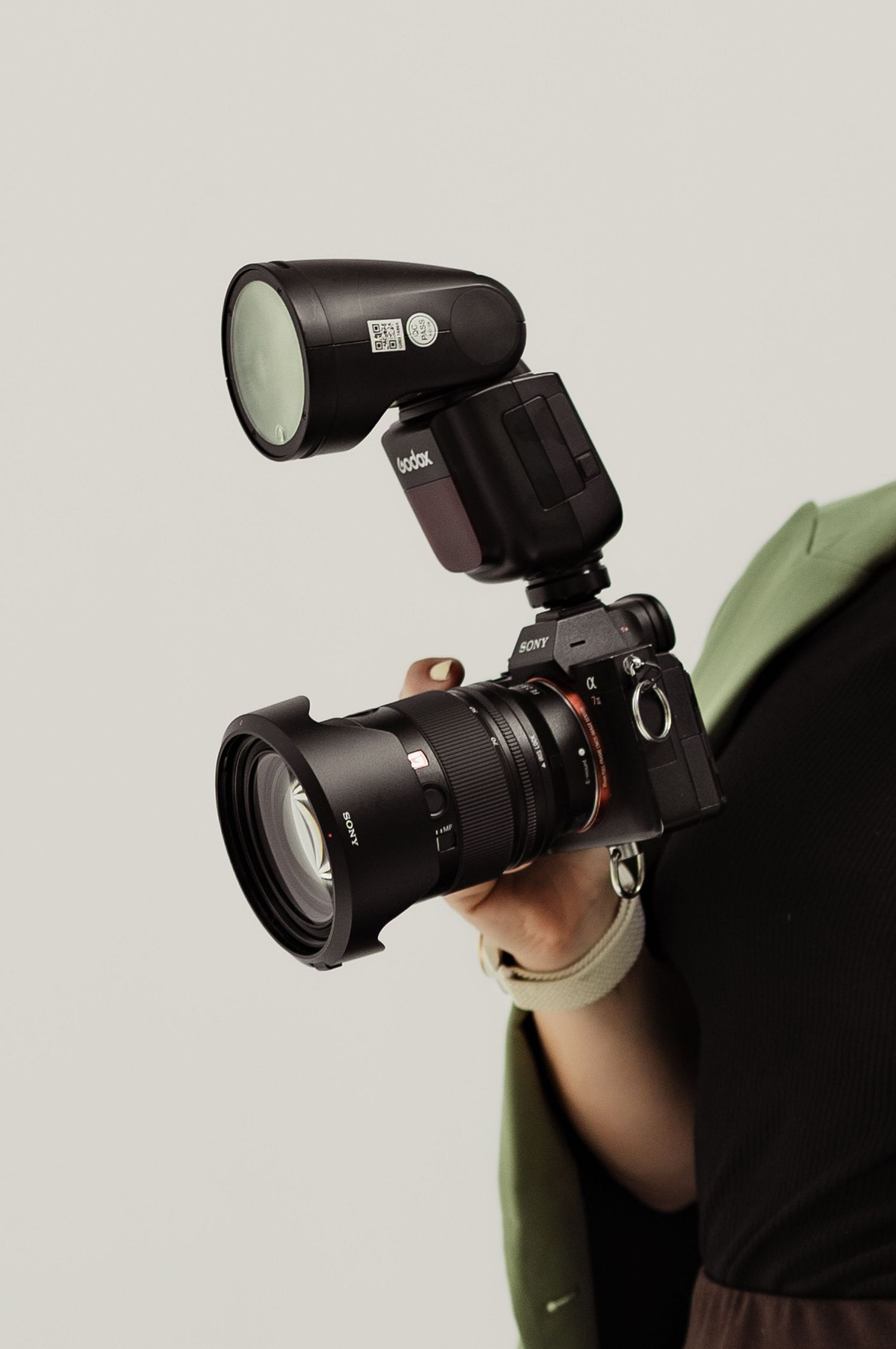 Person holding a Sony mirrorless camera with a Godox flash unit mounted on the hot shoe against a neutral background.