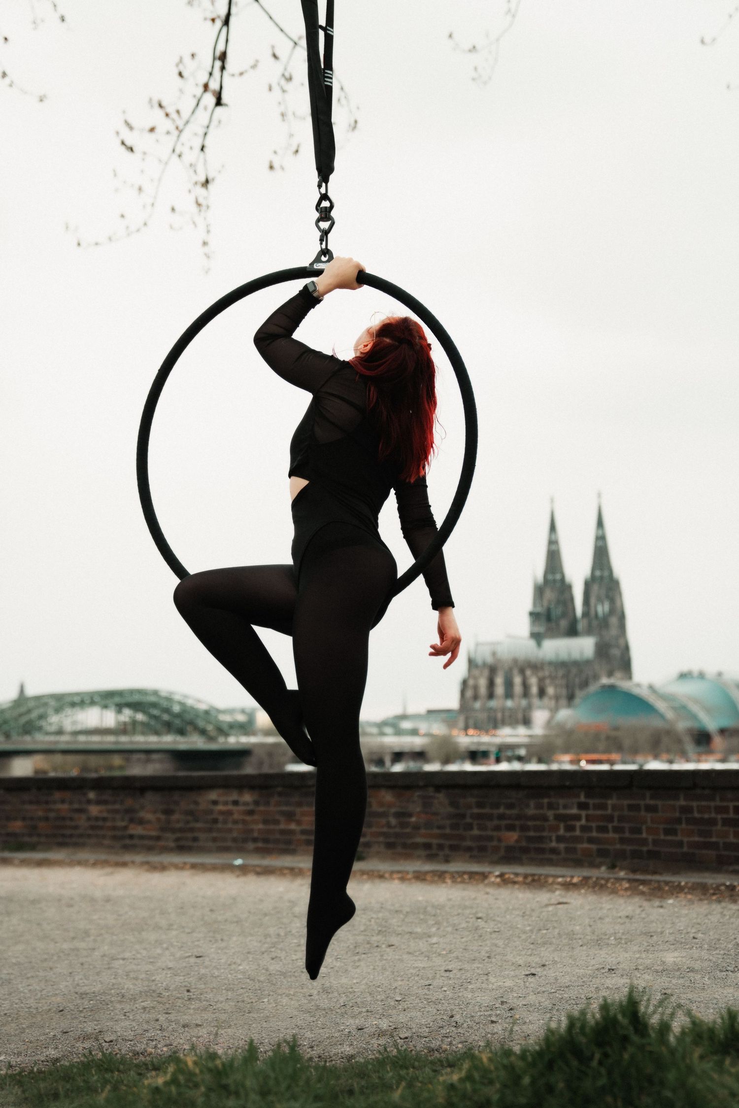 Red-haired aerial hoop performer in black outfit poses outdoors with Cologne Cathedral in background.