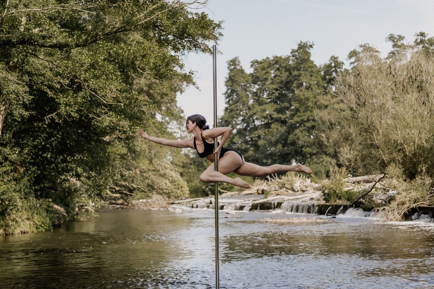 A female pole dancer performs an elegant aerial pose on a pole over a scenic river surrounded by lush green trees.