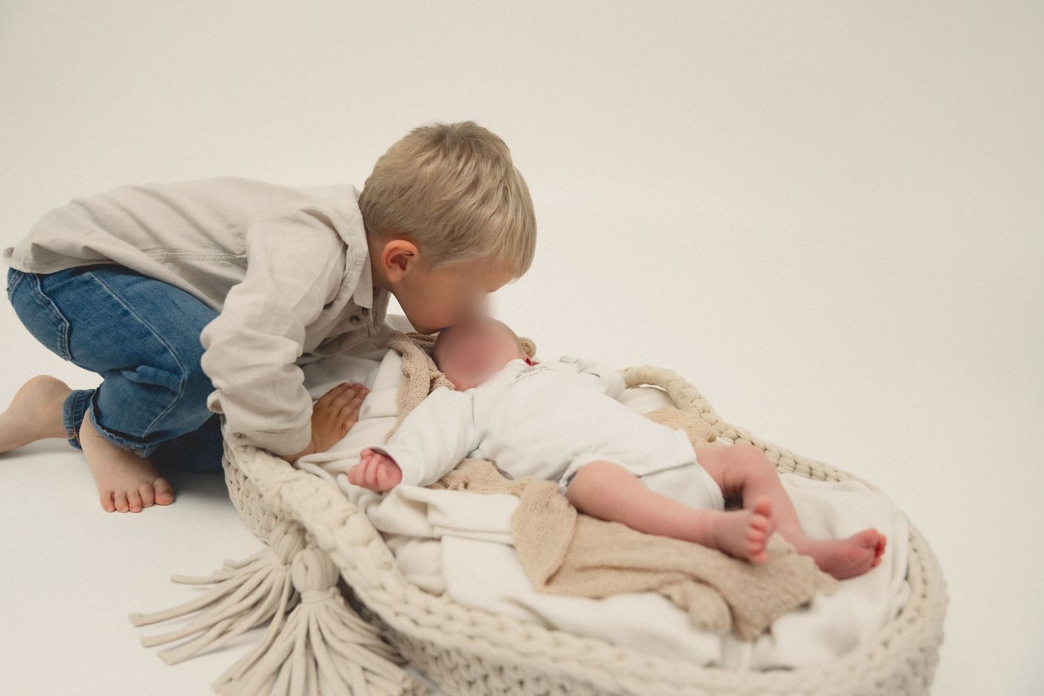 Young boy leaning over to kiss newborn baby sibling lying in a woven Moses basket with cream blanket.
