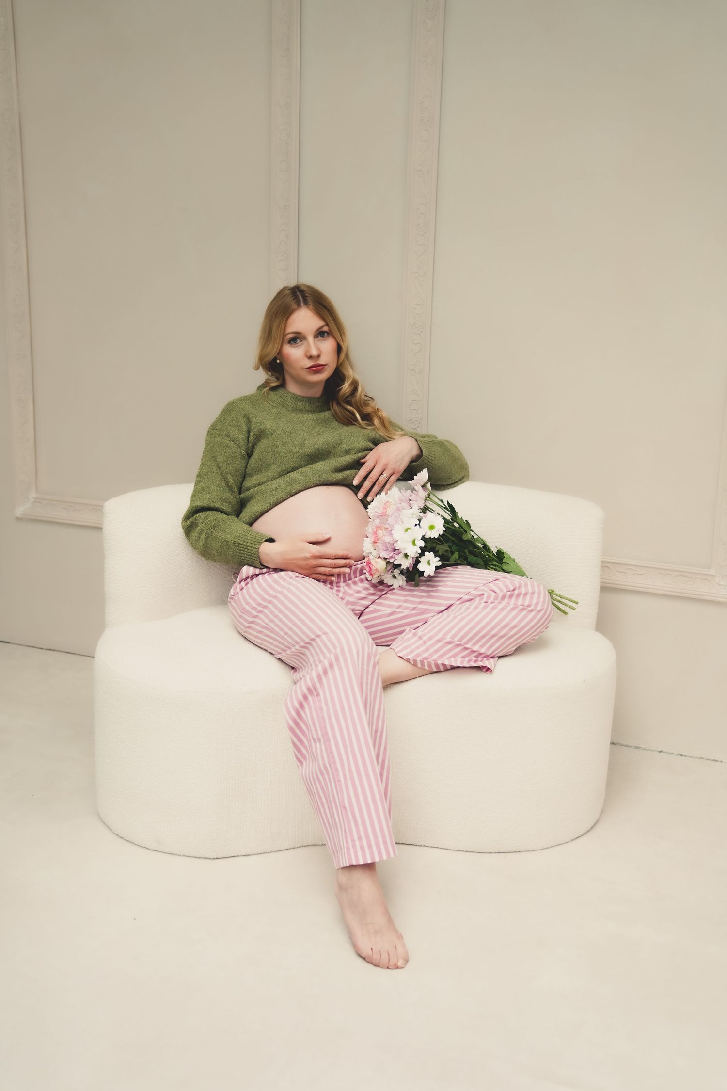 Pregnant woman in green sweater and pink striped pants sitting on white chair holding a bouquet of daisies.