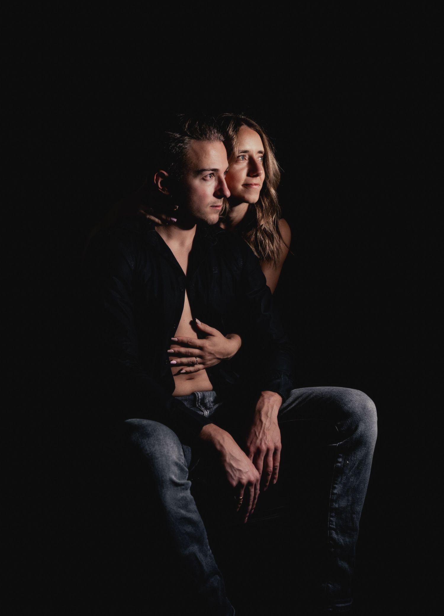 A couple embraces intimately in a dramatic dark studio portrait, wearing black outfits against a black background.