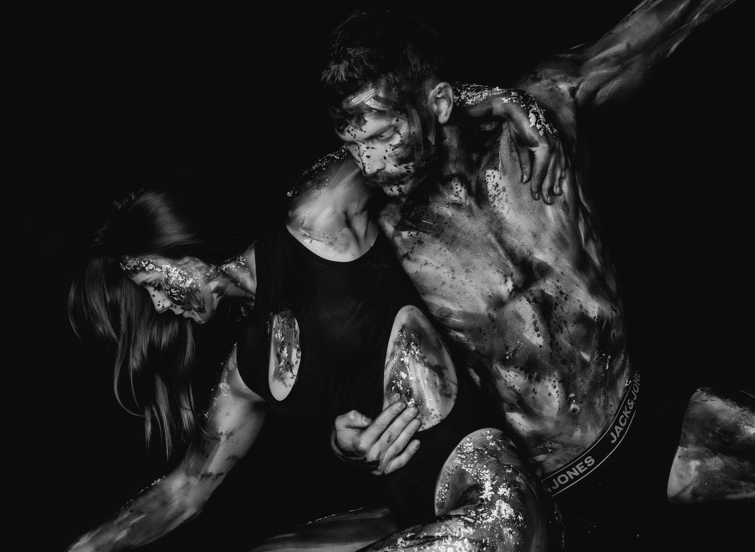 Black and white photo of two muscular figures covered in paint or mud in a dramatic, intense pose.