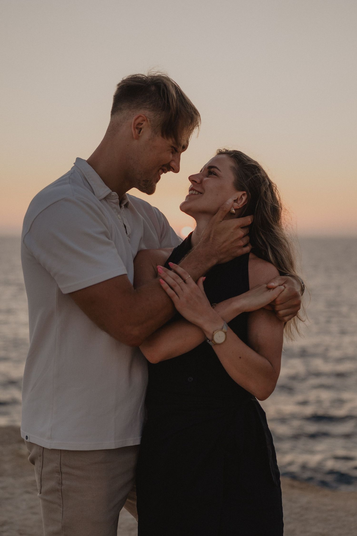 Couple embracing romantically at sunset by the ocean, woman in black dress smiling up at man in white shirt.