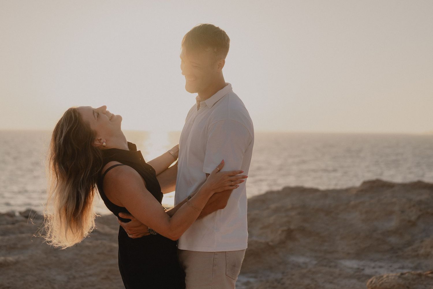 Couple embracing at sunset on rocky coastal cliffs, golden hour light glowing behind them near the ocean.