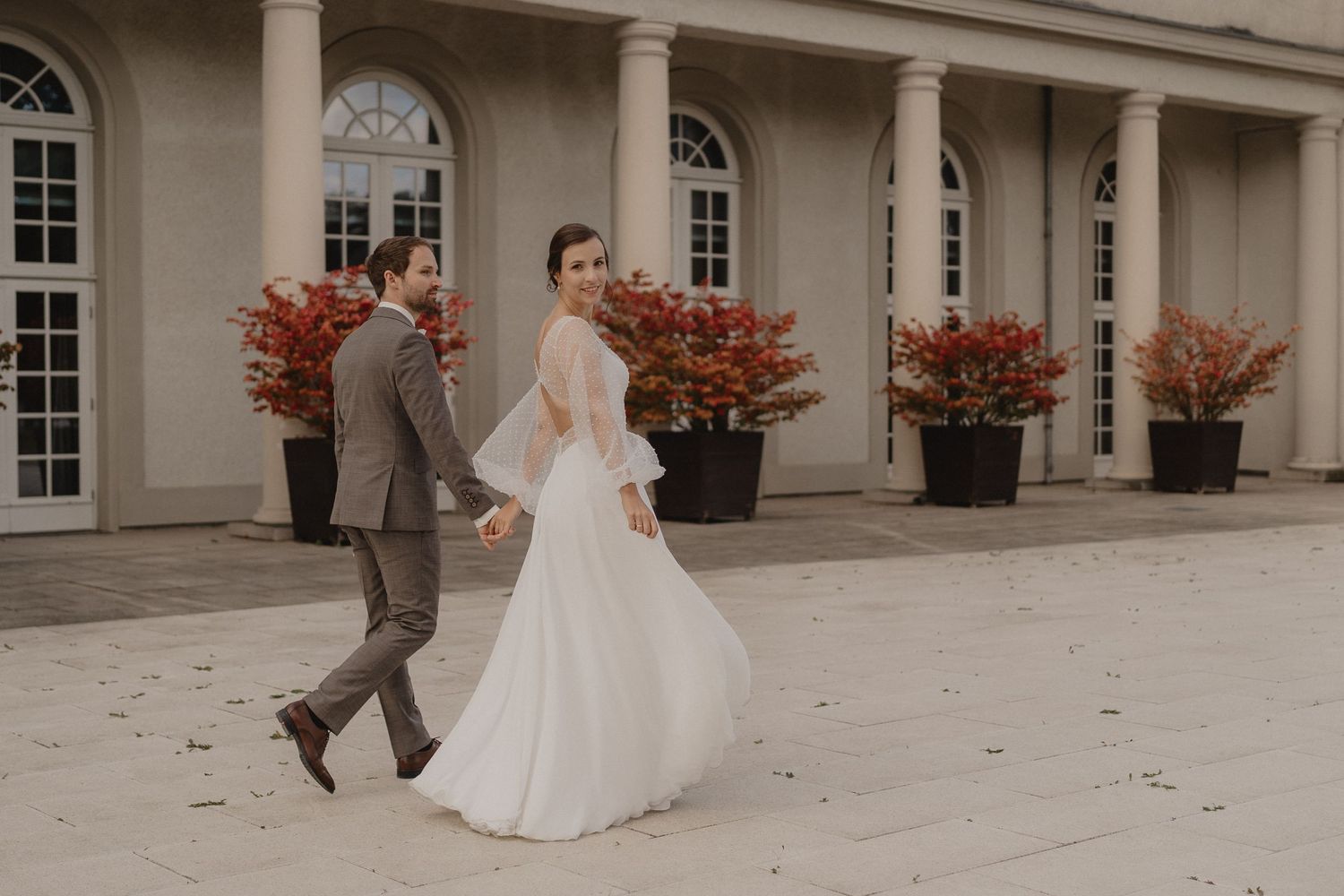A couple in wedding attire walks together outside an elegant building with arched windows and red potted flowers.