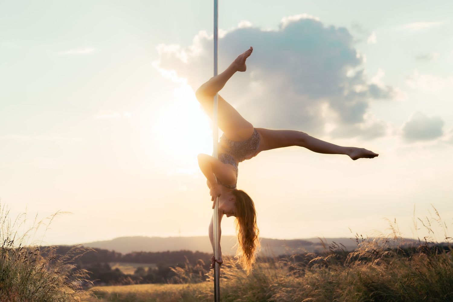 Woman performing a handstand on a pole outdoors at sunset with legs split wide against a dramatic sky.