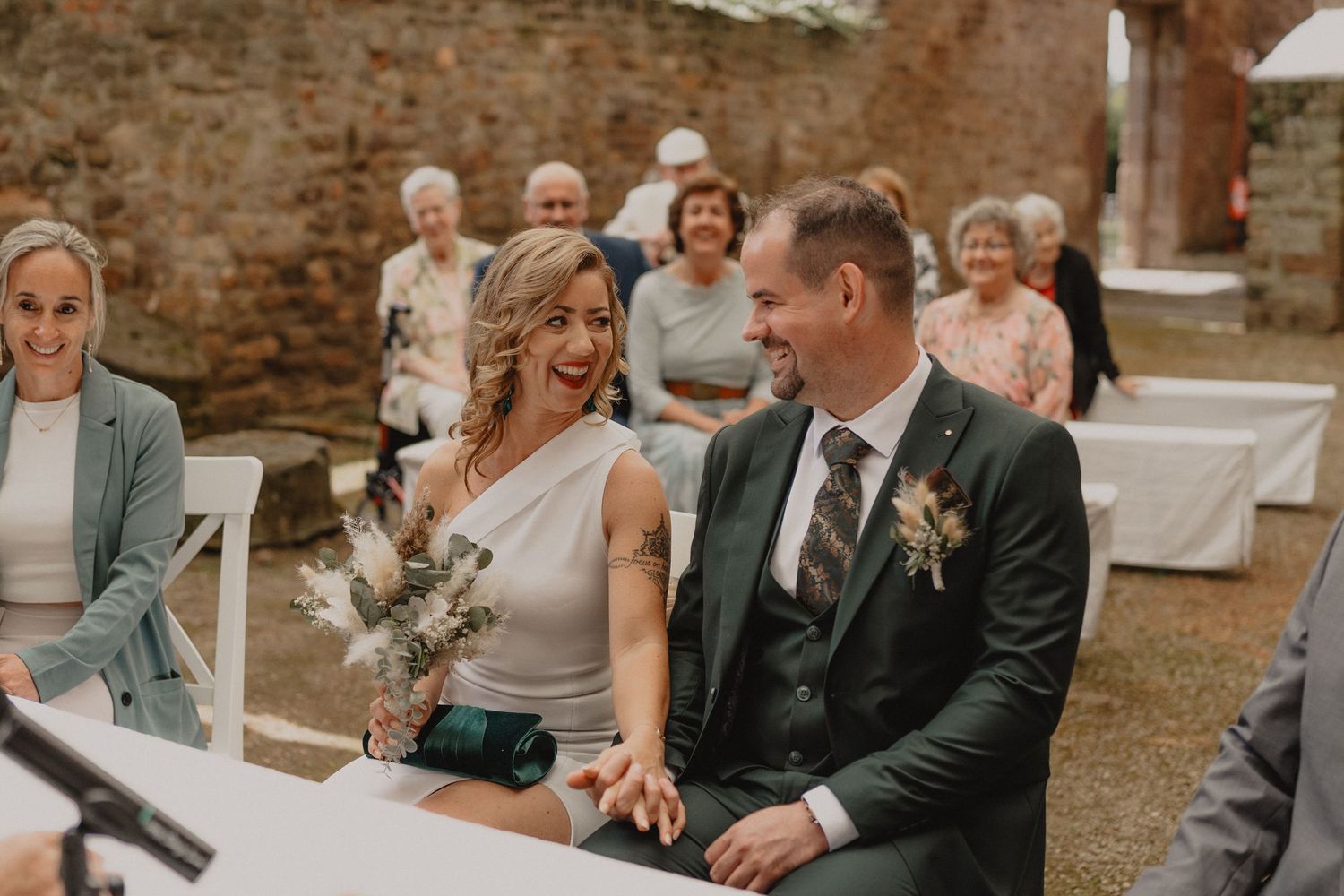 Wedding guests share joyful reactions during an outdoor ceremony at a rustic brick venue.