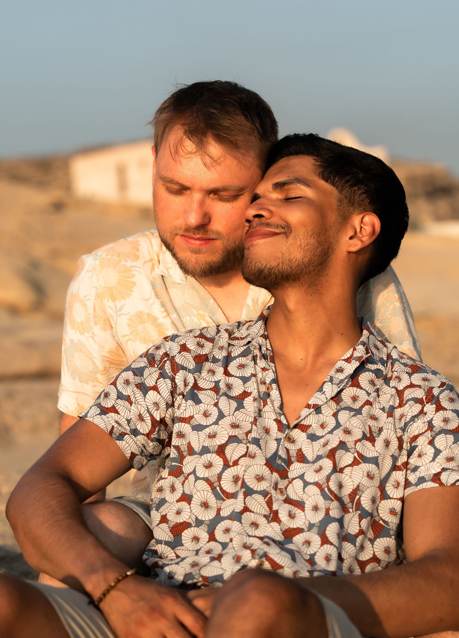 Two young men share a romantic moment outdoors at golden hour, one nuzzling the other's cheek while smiling.