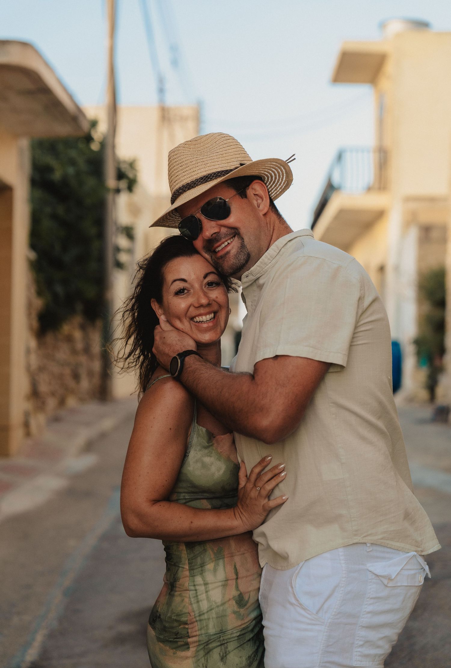 Happy couple embracing on a sunny Mediterranean street, man wearing straw hat and sunglasses, woman in green dress.