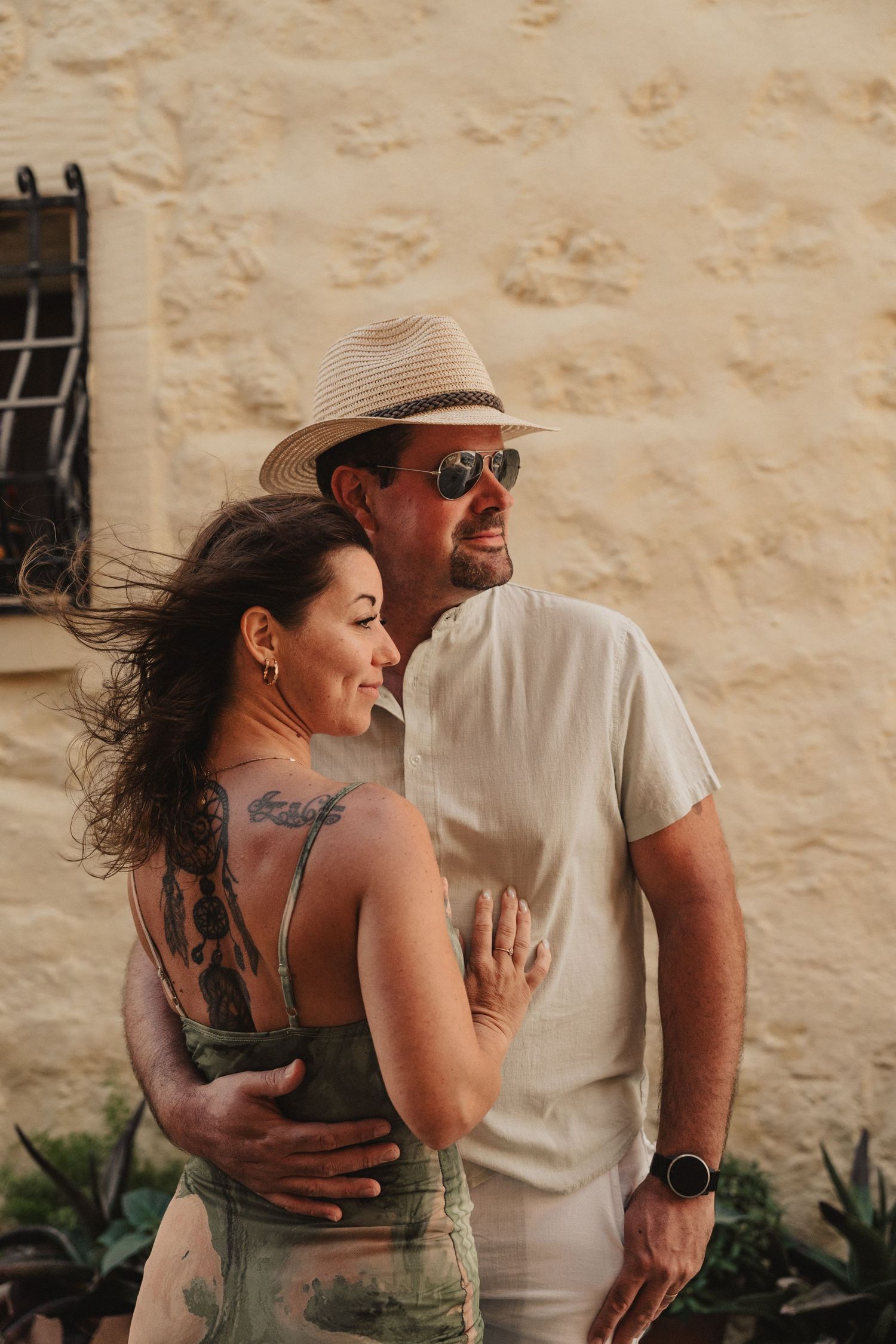A couple embraces outdoors near a stone wall, woman with tattoos in sundress, man in straw hat and sunglasses.