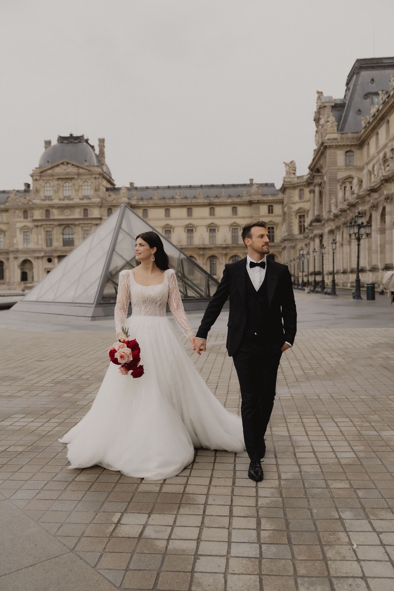 Wedding couple walking hand in hand across a grand courtyard at the Louvre in Paris.