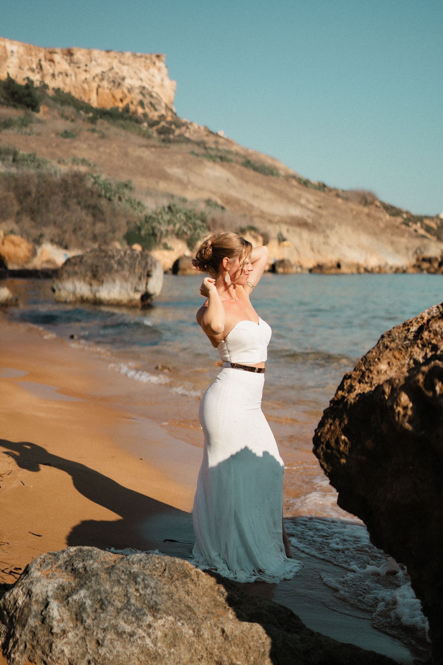 A couple sits together on coastal rocks at sunset, with one wearing a white top and blue skirt against a scenic beach backdrop.