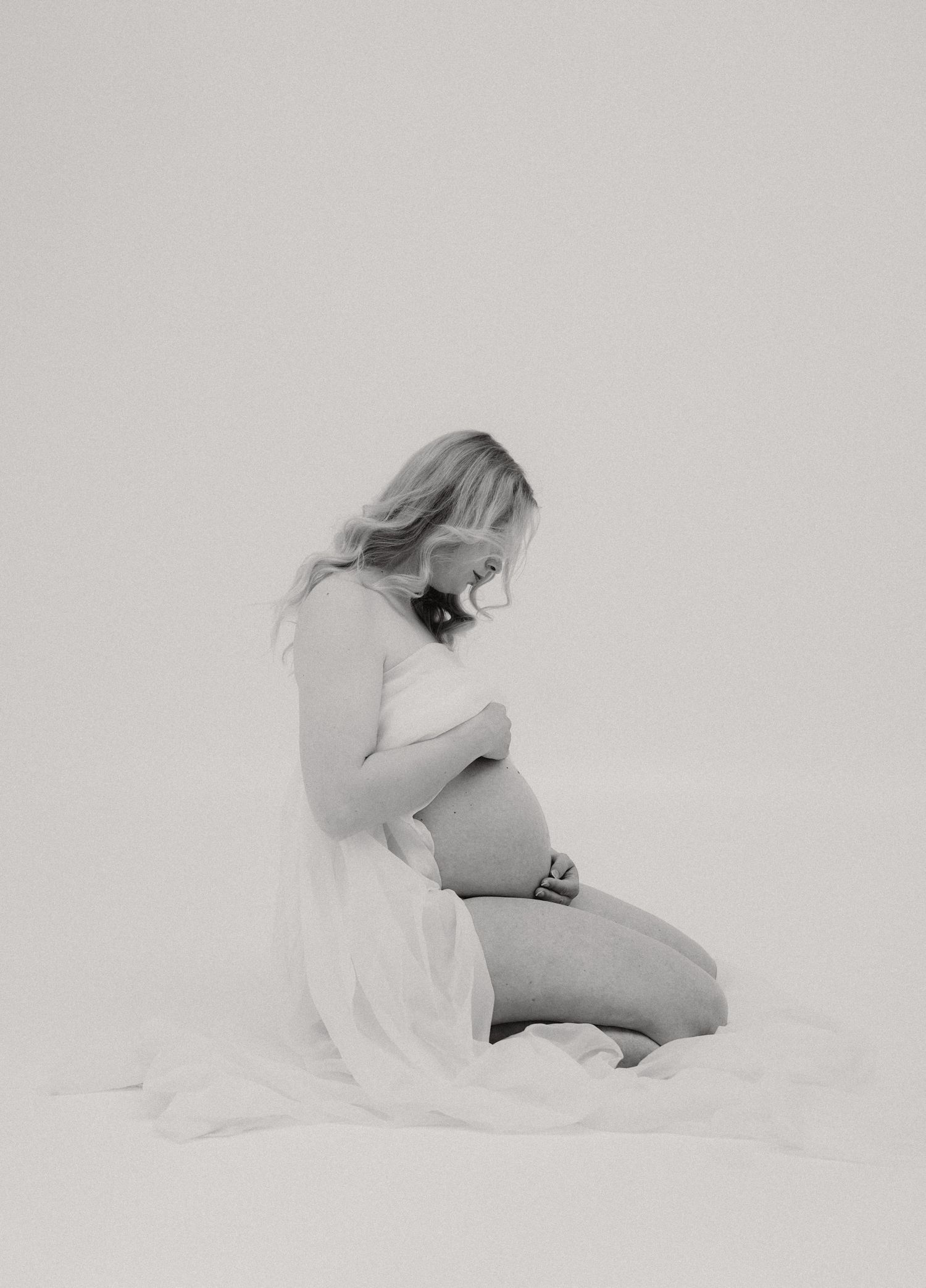 Black and white maternity photo of pregnant woman kneeling gracefully, cradling her belly in a flowing white fabric.
