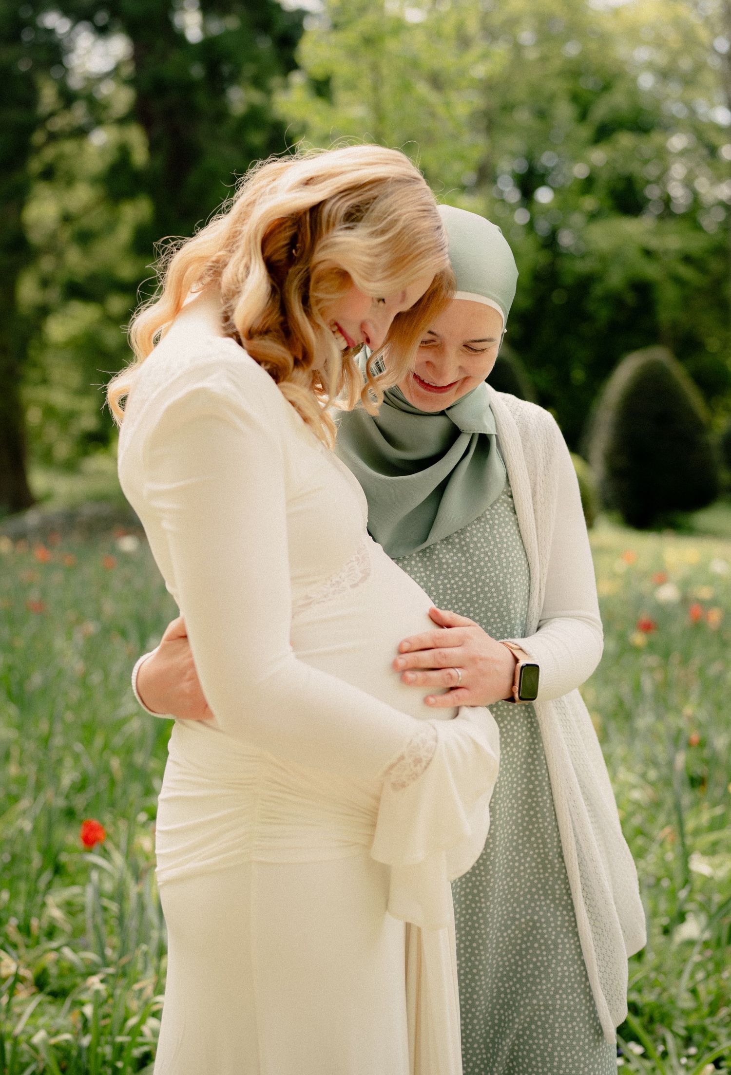 Pregnant woman in white dress embraced by friend in green hijab in a blooming garden.
