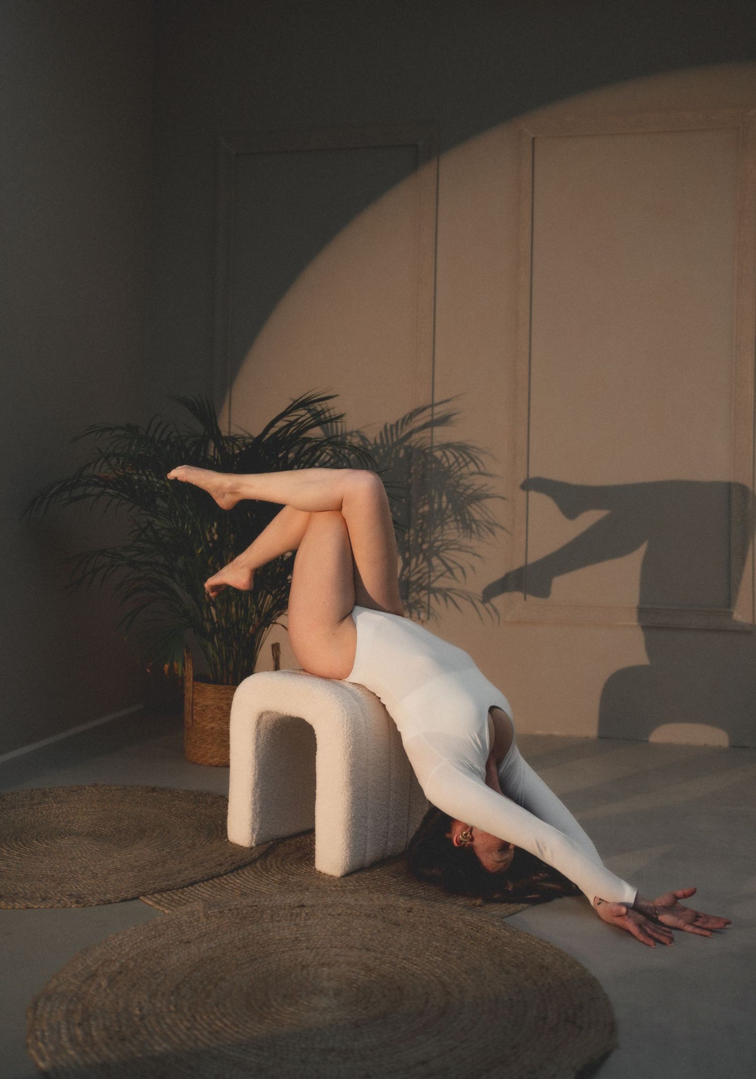 Woman in white bodysuit performing a backbend on a modern stool, with a plant and warm shadows in background.