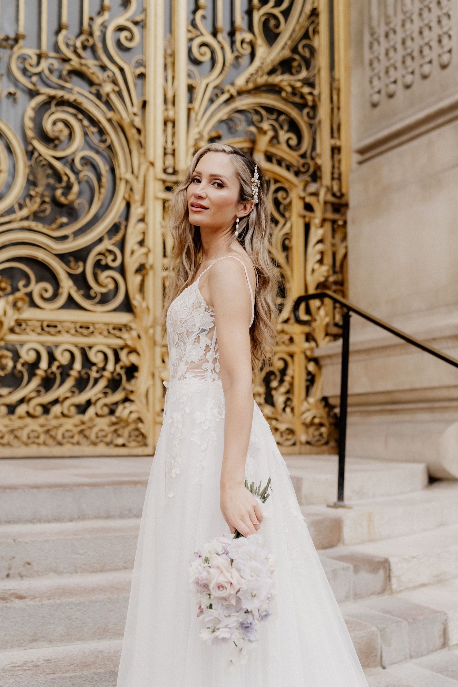Elegant bride in lace white wedding gown poses on stone steps with ornate gold metalwork gate in background.