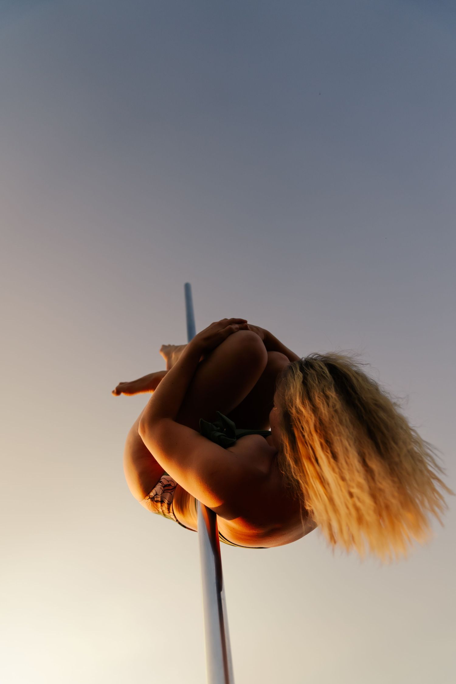 Blonde woman performing an aerial pole dance move against a golden sunset sky.
