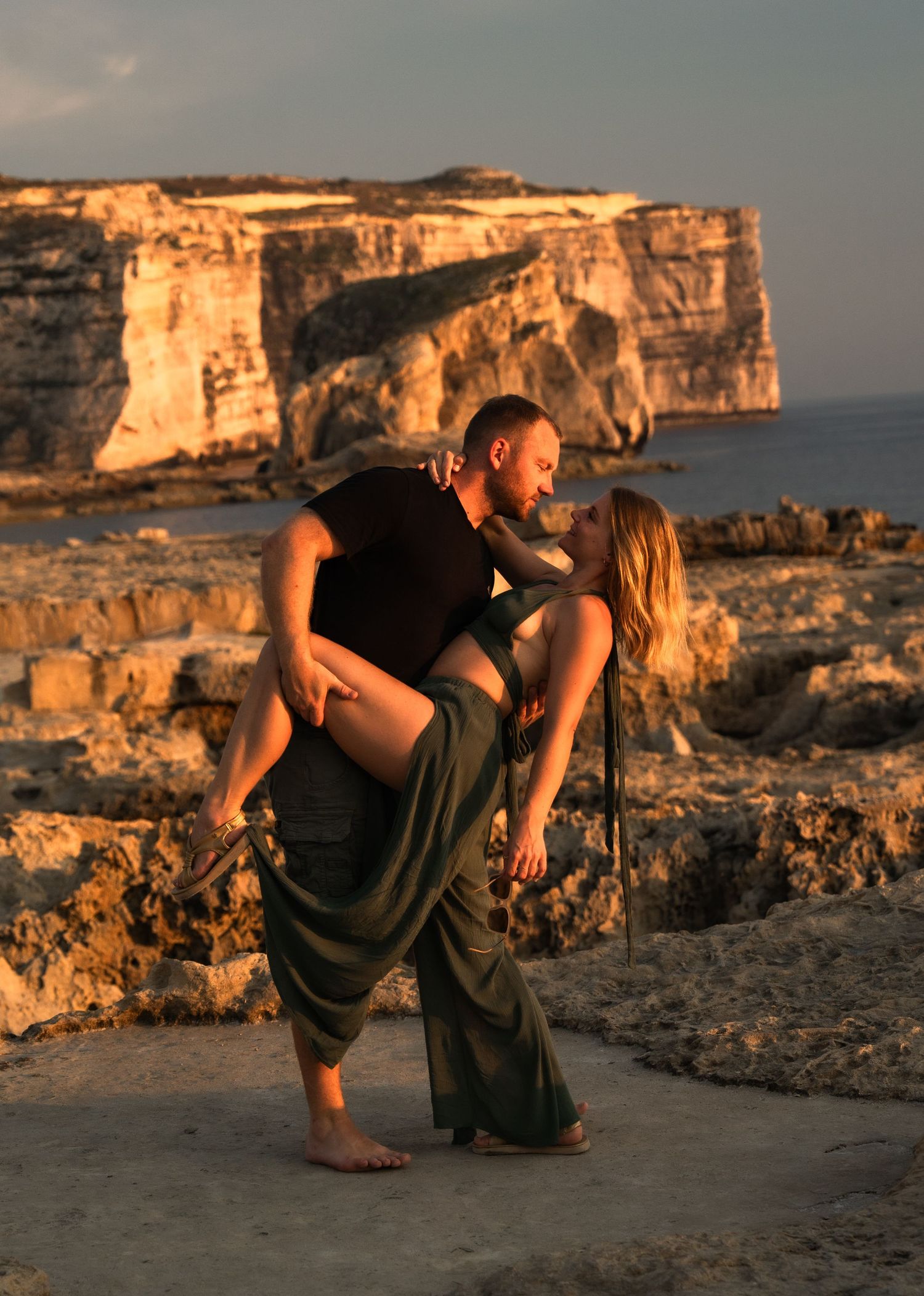 Couple sharing a romantic embrace on rocky coastal cliffs at golden hour sunset.
