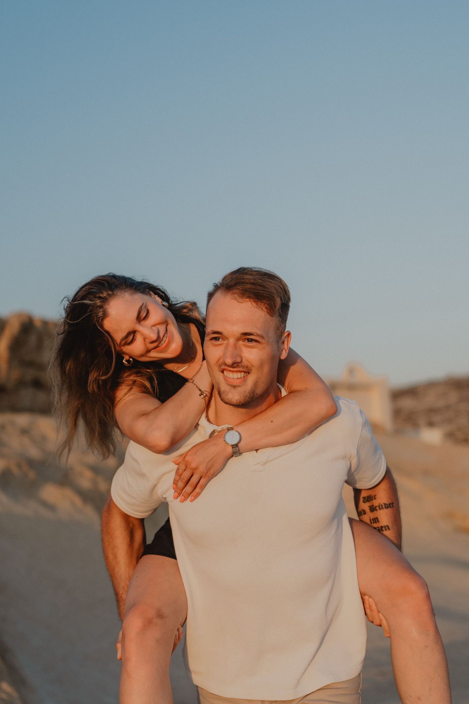Happy couple enjoying a piggyback ride on a sandy beach at sunset, smiling and embracing outdoors.