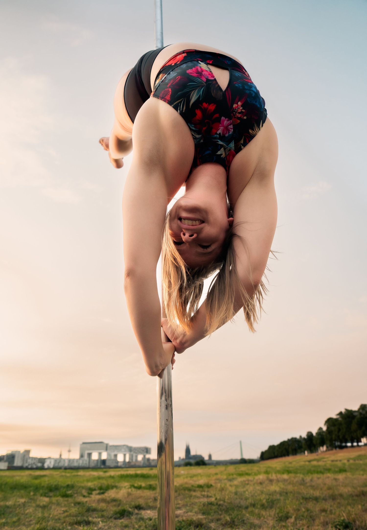 Flexible woman performing an inverted pole dance move outdoors at sunset, wearing a floral bodysuit.