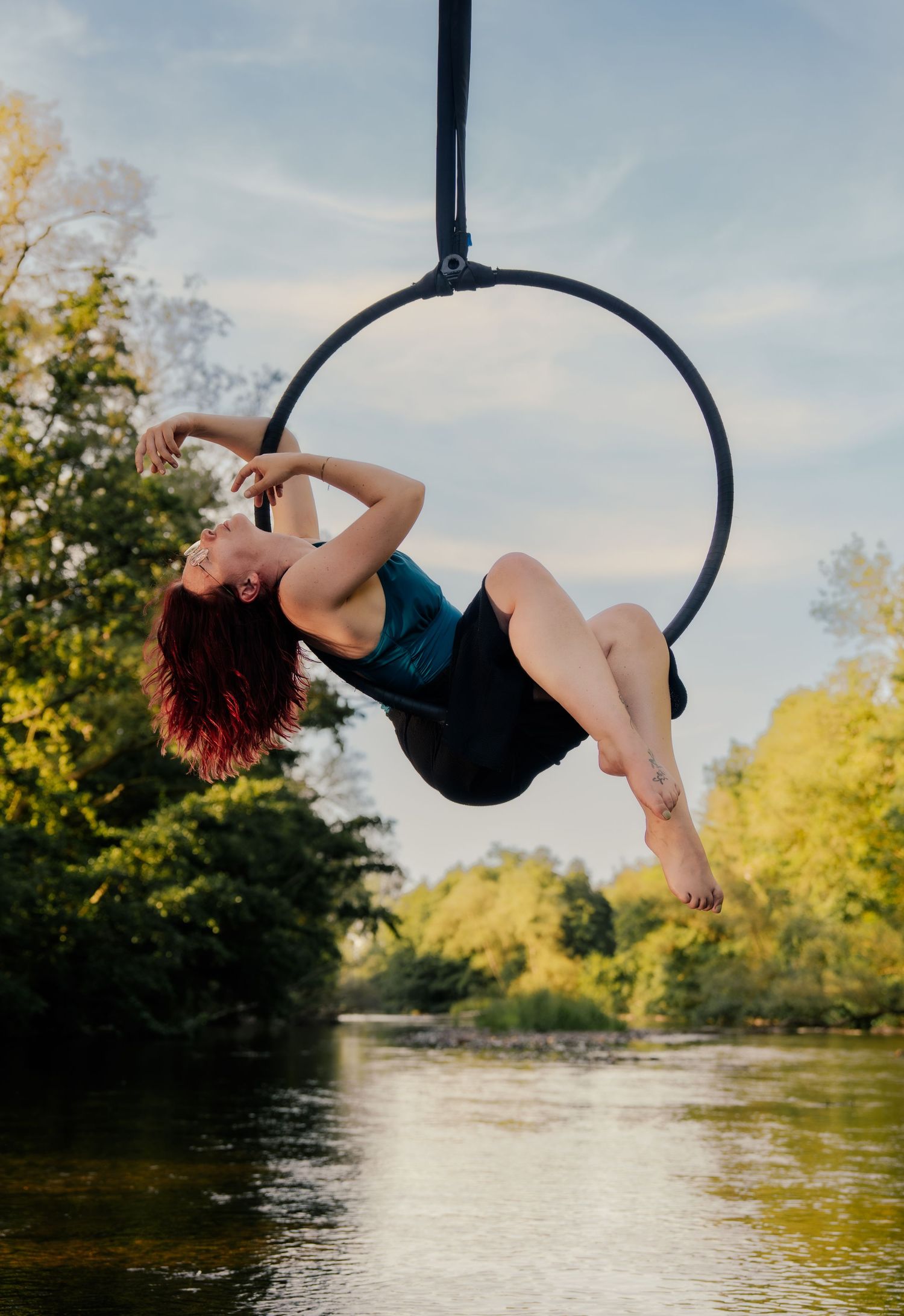 Aerial hoop performer in teal leotard poses gracefully over a river surrounded by lush green trees on a sunny day.