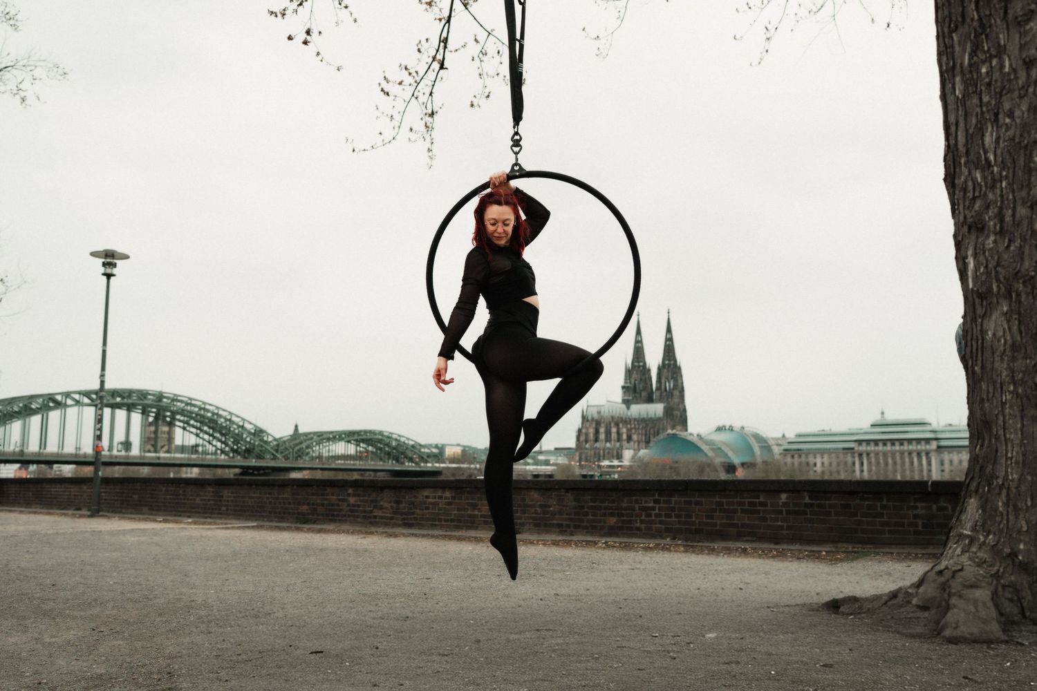 Red-haired aerial hoop artist in black outfit posing gracefully outdoors near Cologne Cathedral.