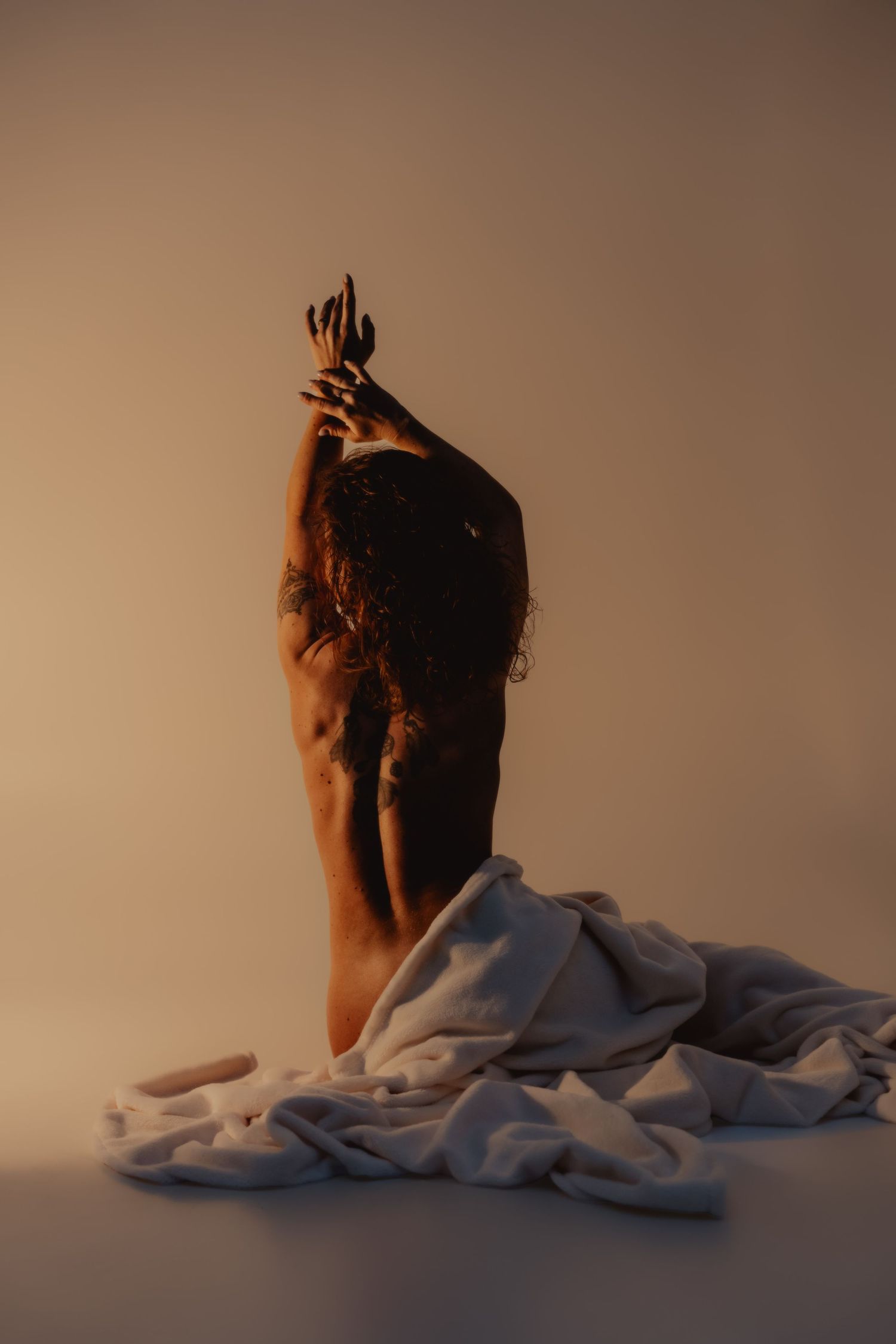 A person sitting on the floor draped in white fabric, arms raised gracefully in warm amber studio lighting.