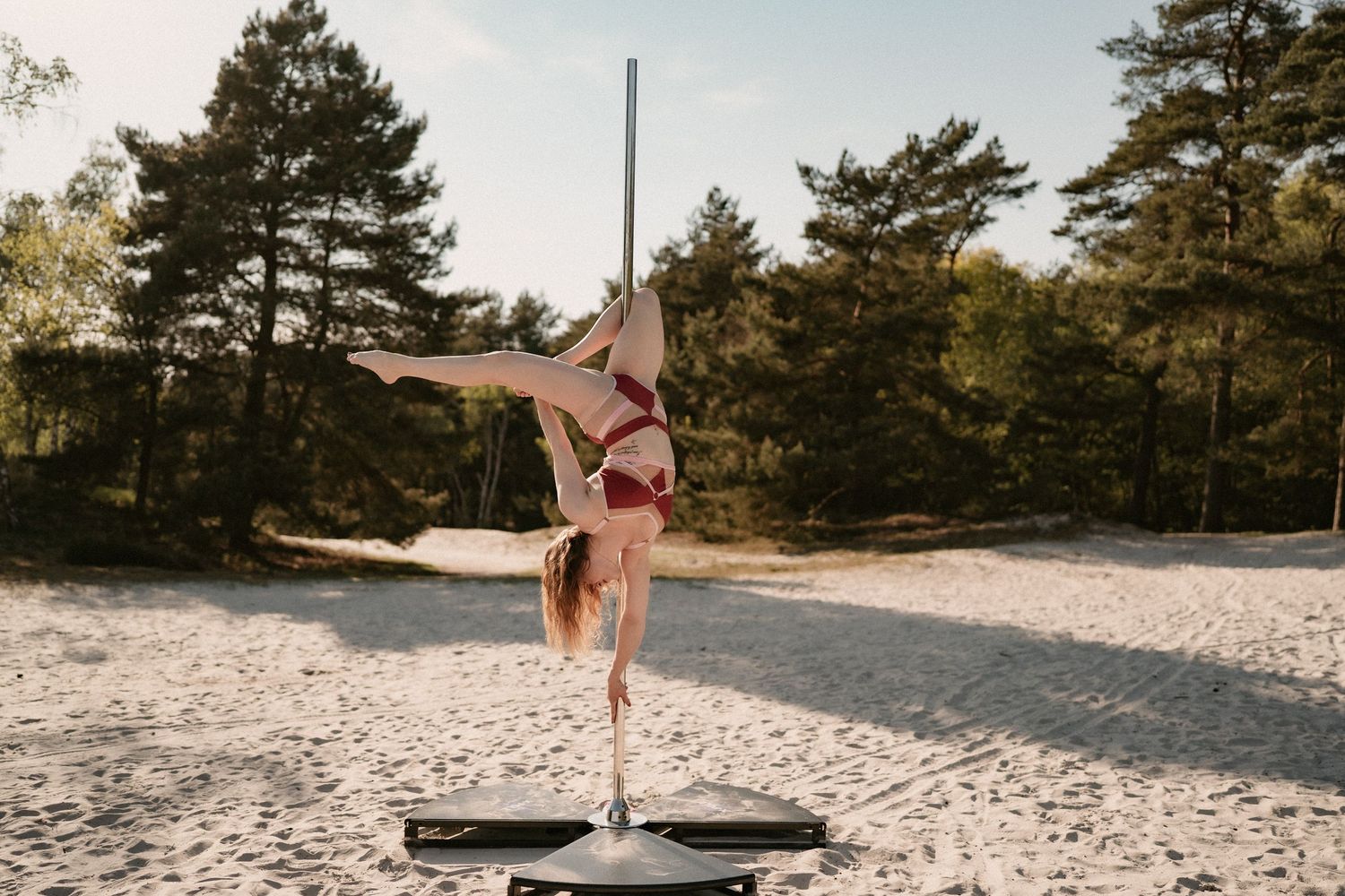 Woman in red bikini performing a handstand on a balance board outdoors on sandy ground with pine trees in background.