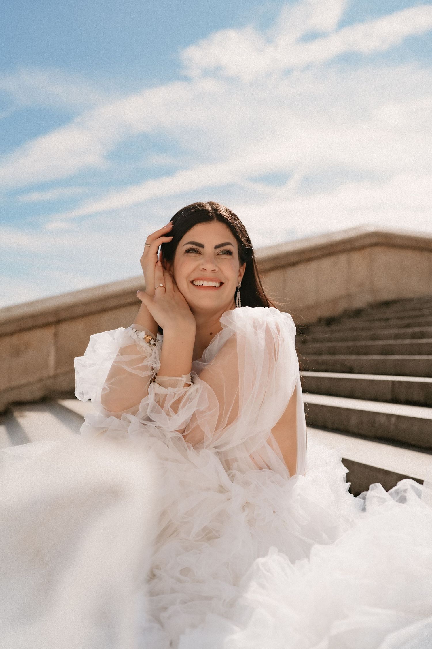 Joyful bride in flowing white dress twirling on stone steps under blue sky.