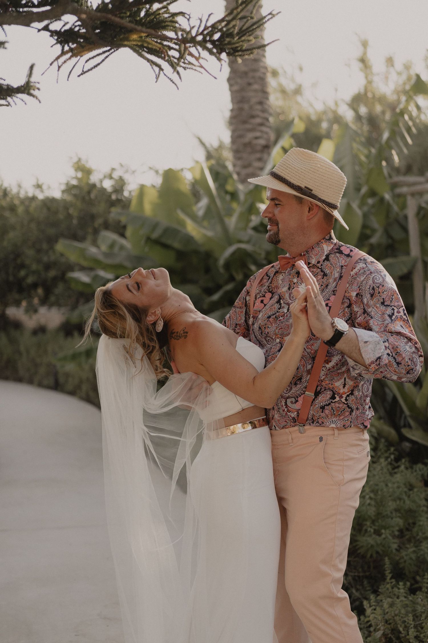 A couple in wedding attire share a joyful dance moment in a tropical garden setting.