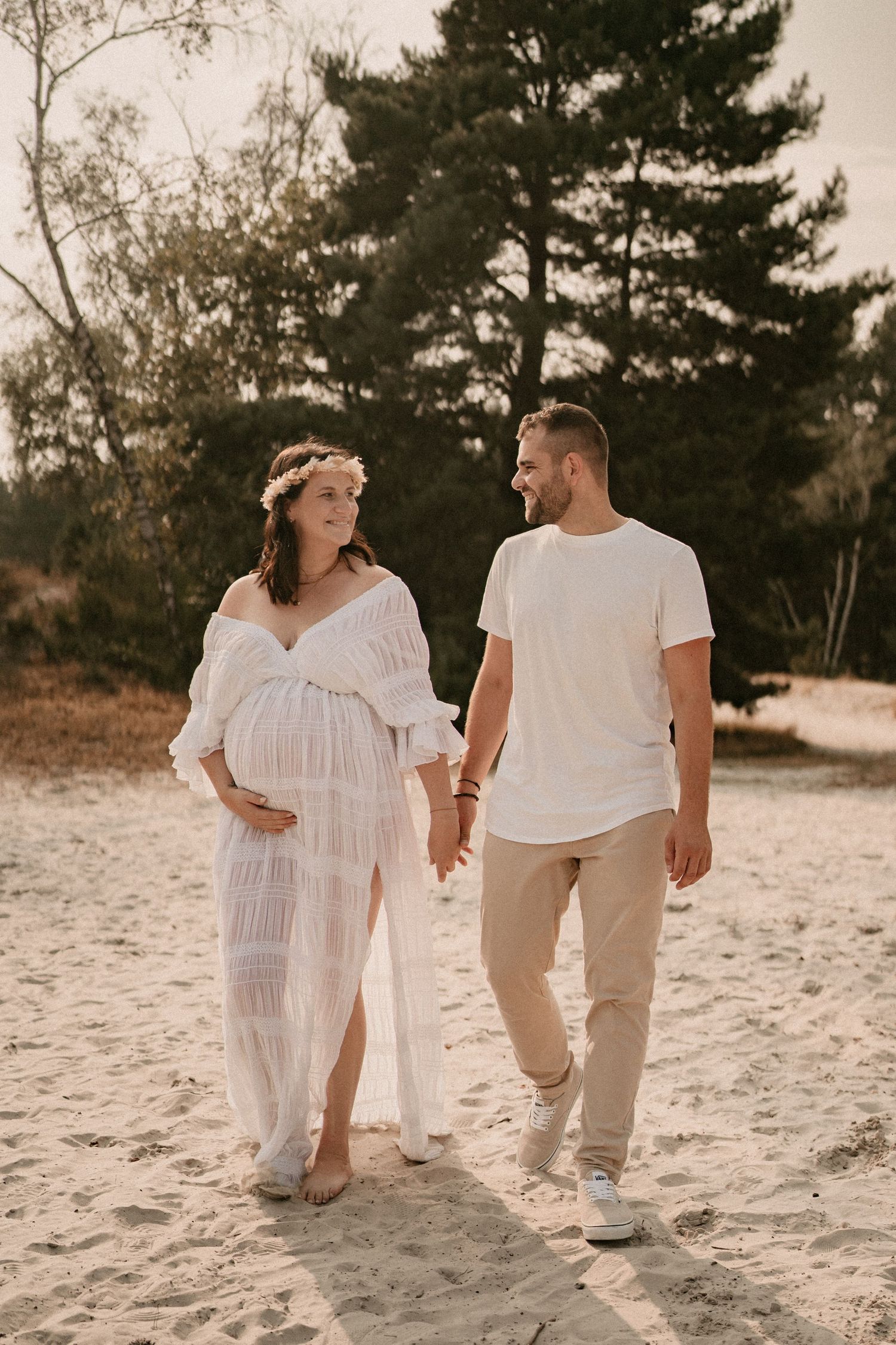A couple walks hand in hand on a sandy beach during a maternity photoshoot.