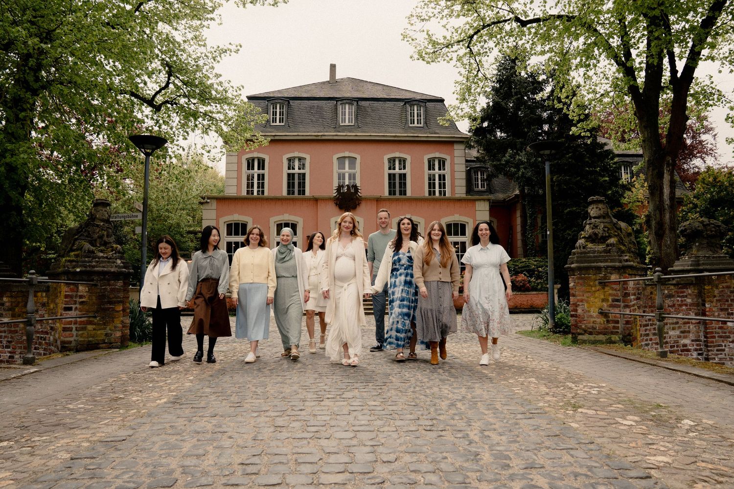 Group of elegantly dressed women walking on cobblestone path before a pink manor house.