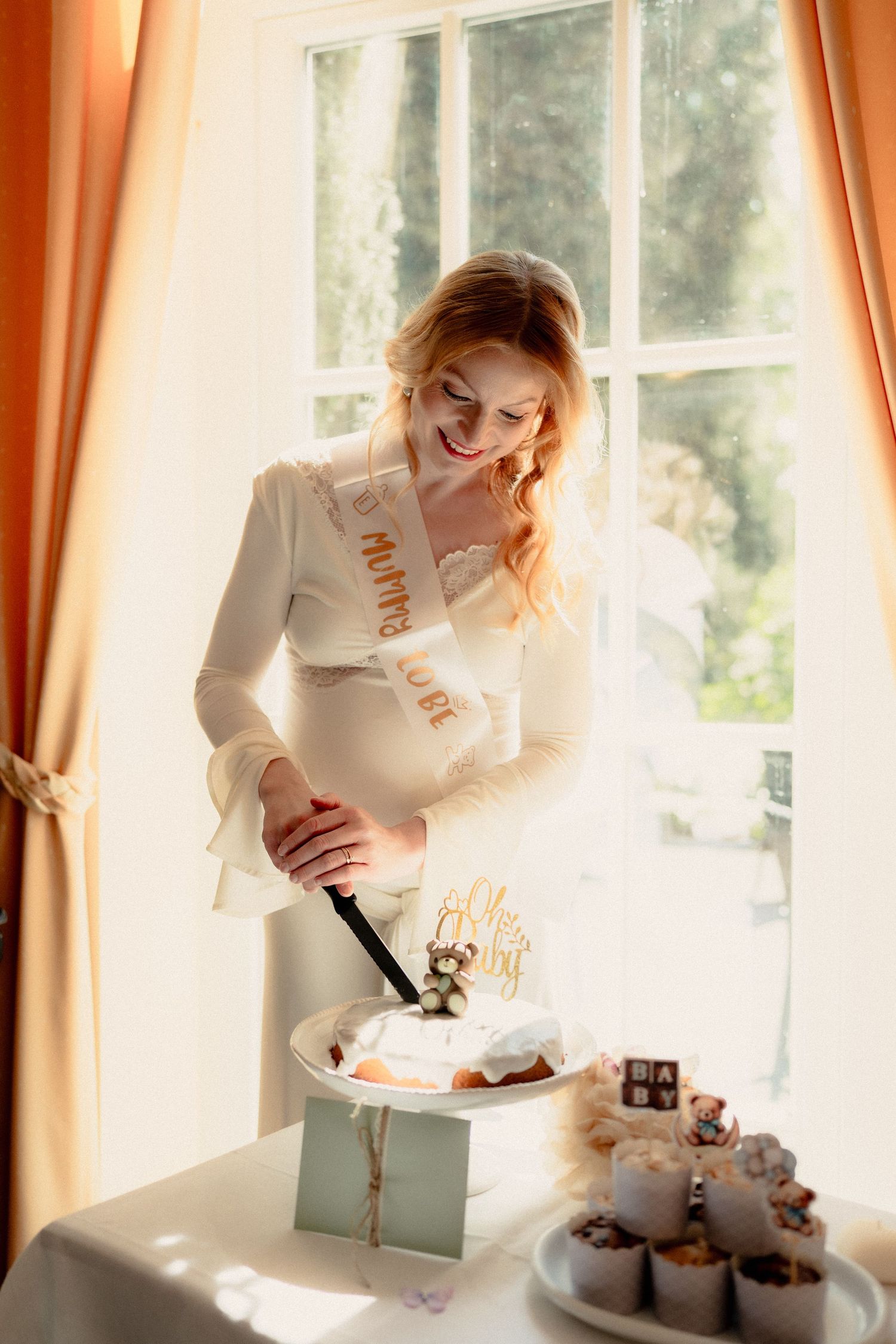A smiling pregnant woman in white cuts a cake at her baby shower near a sunlit window.