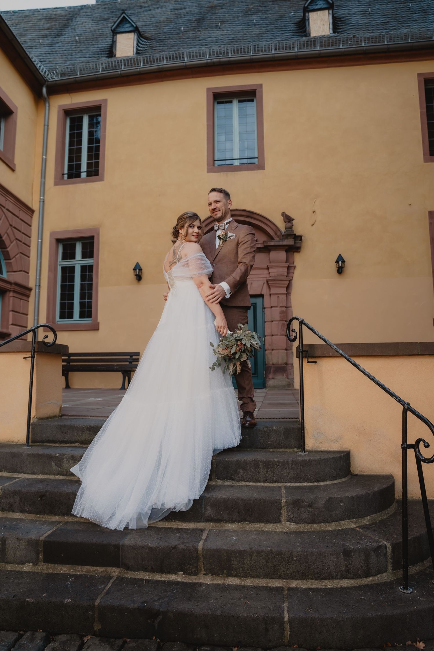 Wedding couple descending stone steps outside a historic yellow building with dark slate roof.