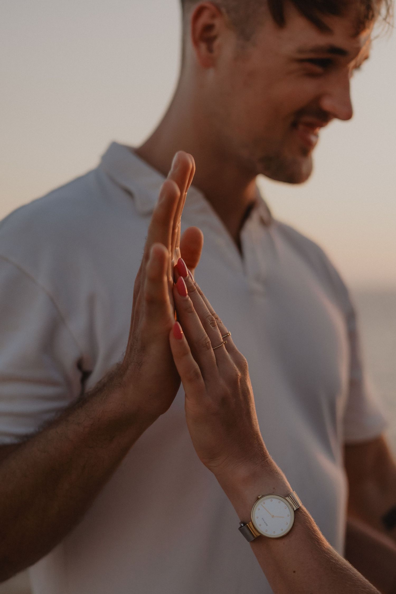 A smiling man in a white polo shirt touching hands with a woman wearing a watch at sunset.
