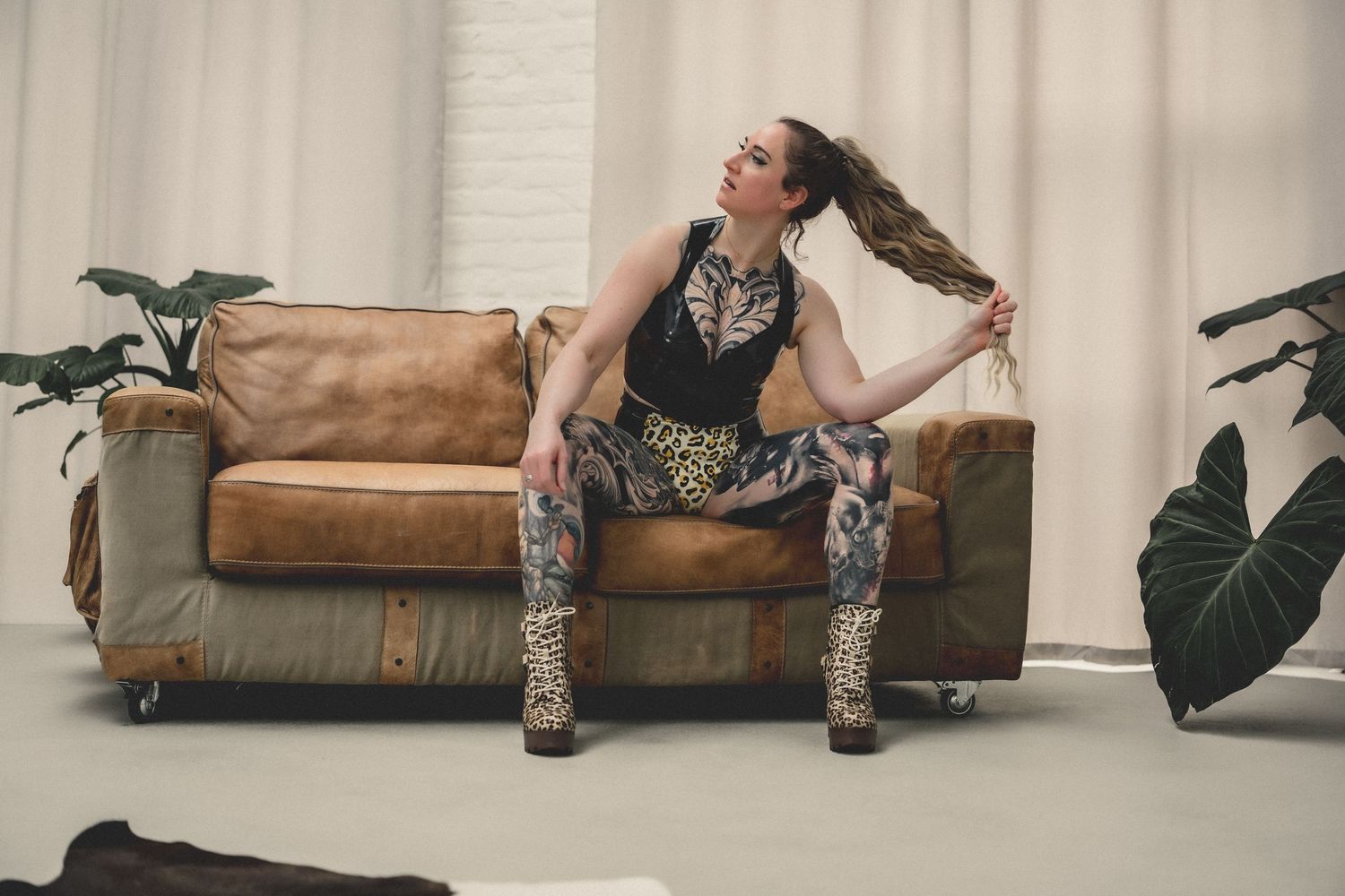 Tattooed woman in leopard print outfit sitting on vintage leather sofa, pulling her ponytail in a bright studio setting.