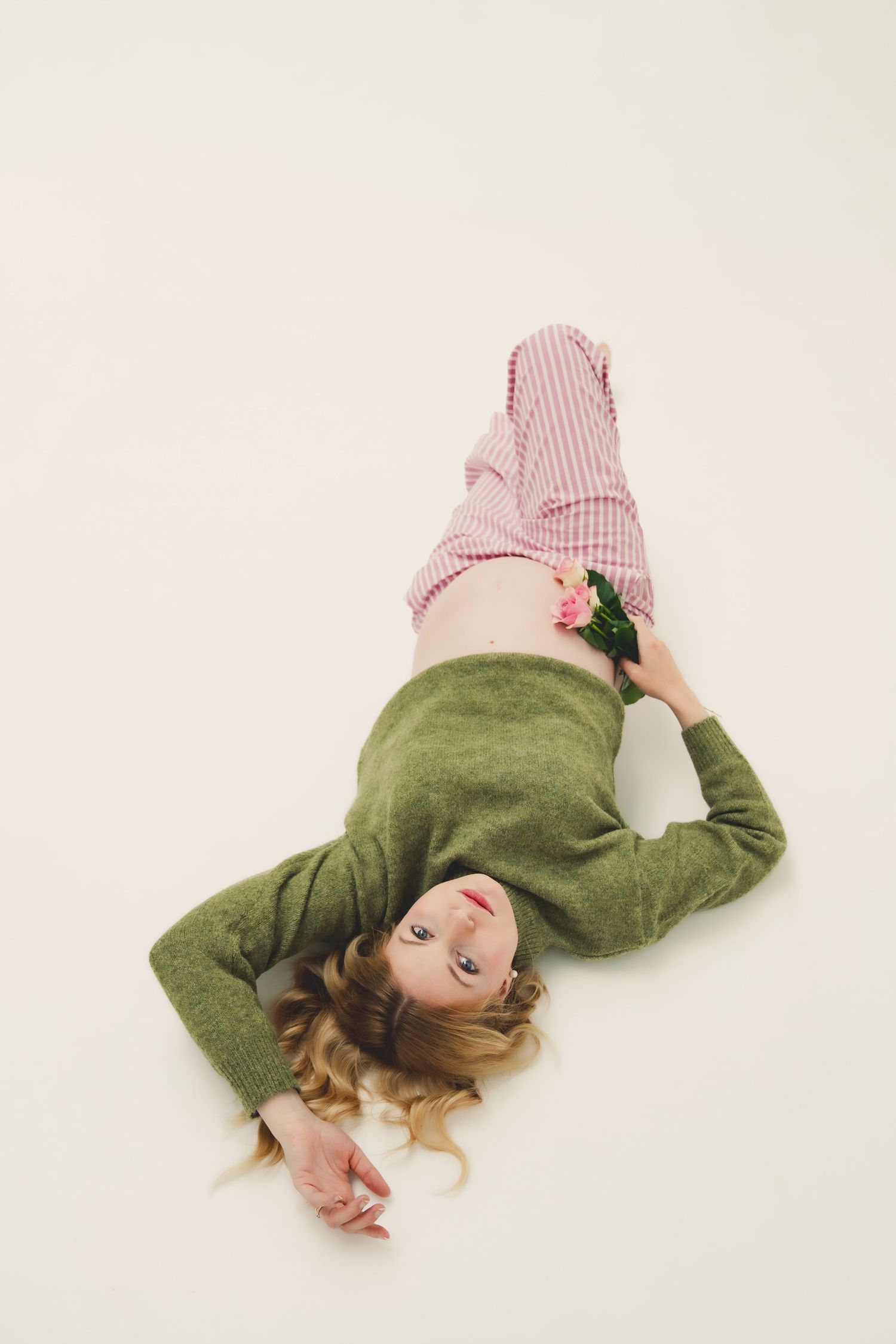 Pregnant woman lying on white floor in green sweater and pink pants, holding pink flowers on her baby bump.