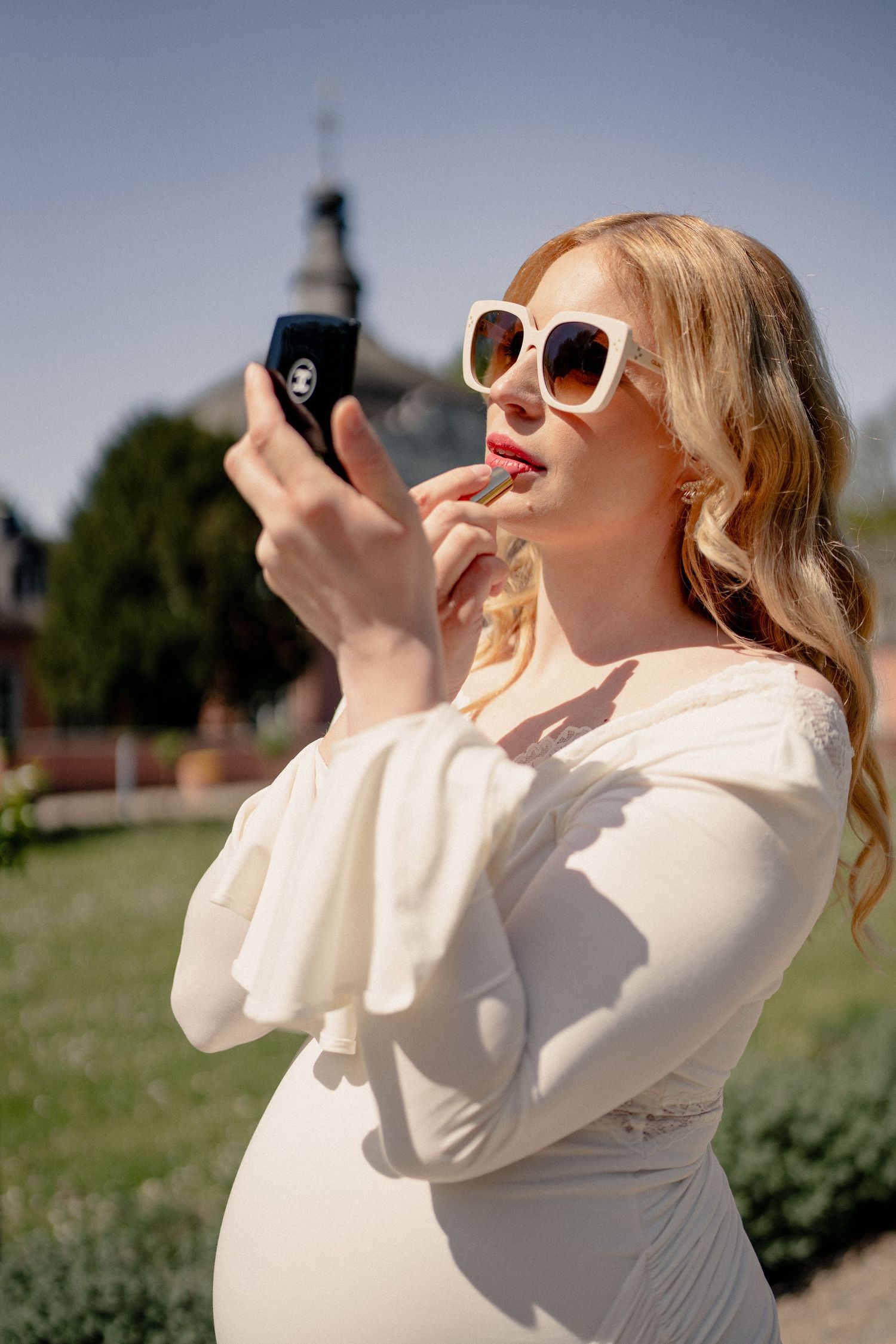 Pregnant woman in cream dress applying lipstick using a compact mirror outdoors on a sunny day.