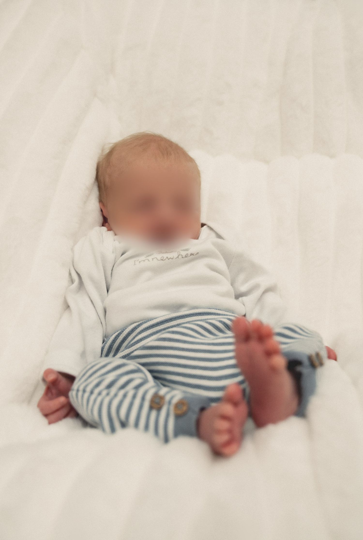Newborn baby lying on white fluffy blanket wearing white bodysuit and blue striped pants with bare feet.