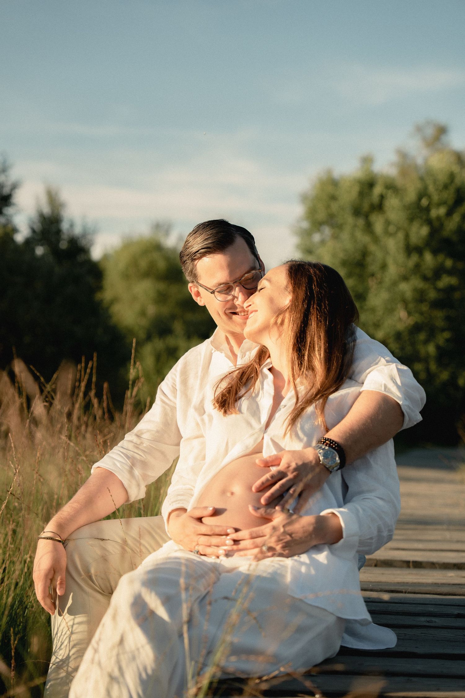 A couple sits on a wooden dock at sunset, the man kissing his pregnant partner as they both cradle her baby bump.
