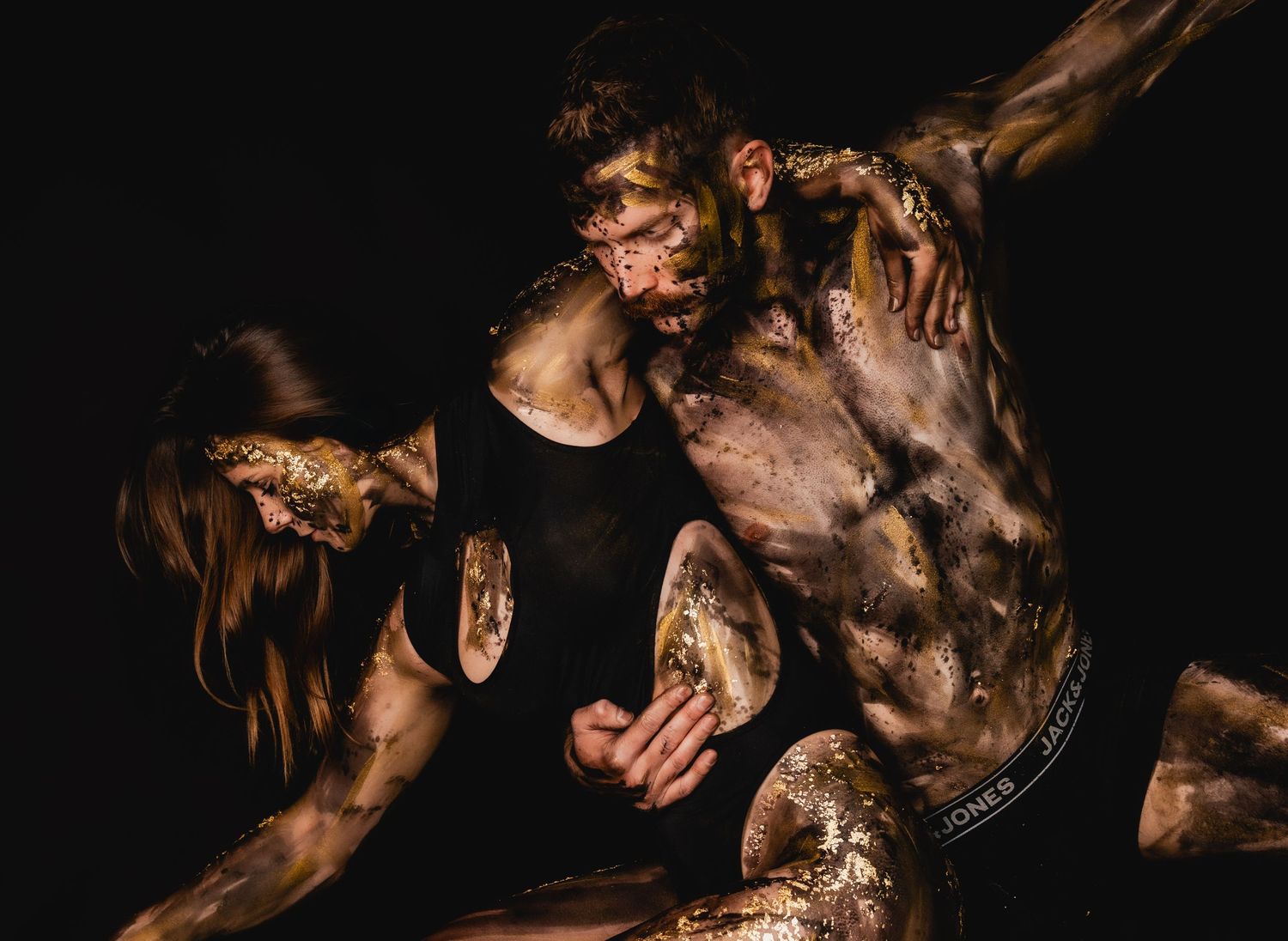 Two performers covered in gold and mud paint pose dramatically against a dark background in an artistic dance performance.